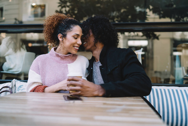 Cheerful diverse young man and woman in casual clothes flirting with eyes closed on date while sitting at table with coffee in outdoor cafe - Photo, image