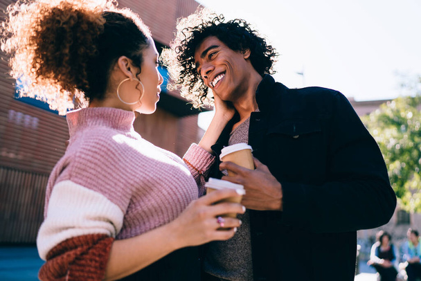 Low angle of young woman touching face of happy African American boyfriend while spending time together and drinking coffee from disposable cups on sunny day - Fotó, kép