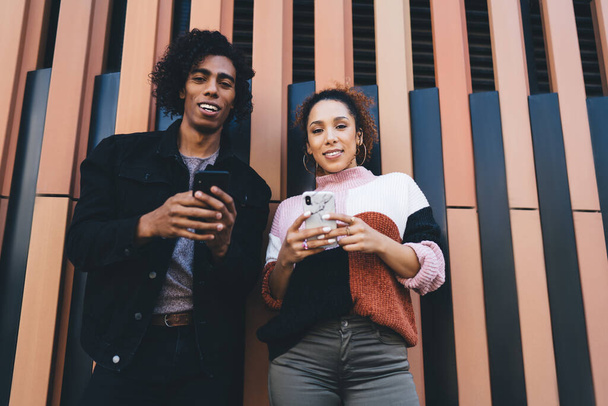 Low angle of positive young African American couple in casual outfits smiling and looking at camera while browsing smartphones leaning on modern wall - Fotó, kép