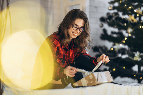 Christmas, New Year. Pretty woman in shirt and socks lying in bed with christmas gift box, new year tree behin - Фото, изображение