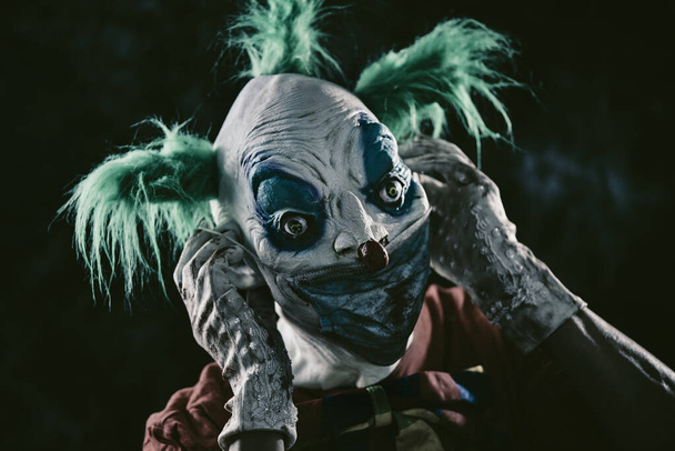 closeup of a disturbing evil clown, with green hair, wearing a dirty red costume, a dirty bow tie, and dirty and bloody gloves, putting on or taking off a nasty face mask, against a dark background - Photo, Image