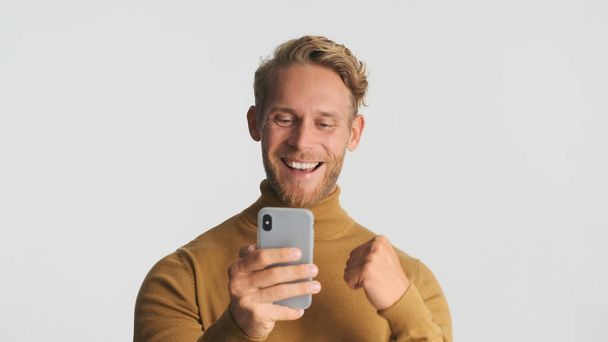 Knappe blonde baardman blij over goede zakelijke deal met behulp van smartphone voor het werk geïsoleerd op witte achtergrond - Foto, afbeelding