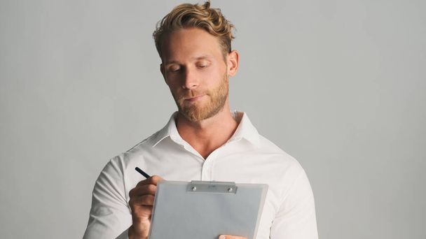 Красивый концентрированный белокурый бородатый бизнесмен в рубашке, пристально работающий с документами на белом фоне. Концепция бизнеса - Фото, изображение