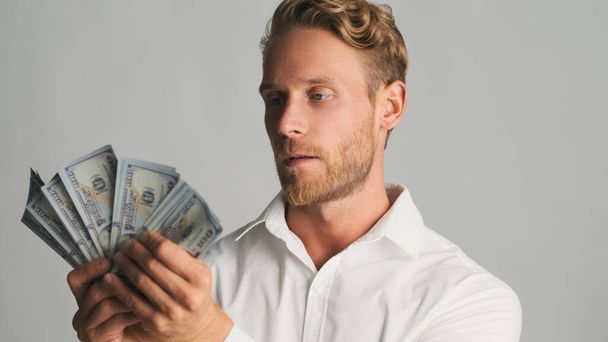 Handsome blond bearded businessman looking serious posing with money on camera over white background - Foto, imagen