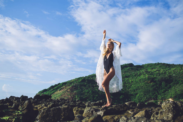 Full body slim female in swimwear raising arms and closing eyes while standing on stones of green hill against cloudy sky on sunny day - Fotografie, Obrázek