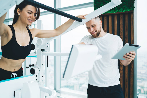 Cheerful slim sportswoman in activewear training on sports machine and smiling while having workout in modern light gym with trainer - Фото, изображение