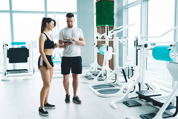 Fit female with dark hair in sportswear having conversation with male instructor browsing tablet in urban spacious gym with modern exercise machines - Foto, Imagen