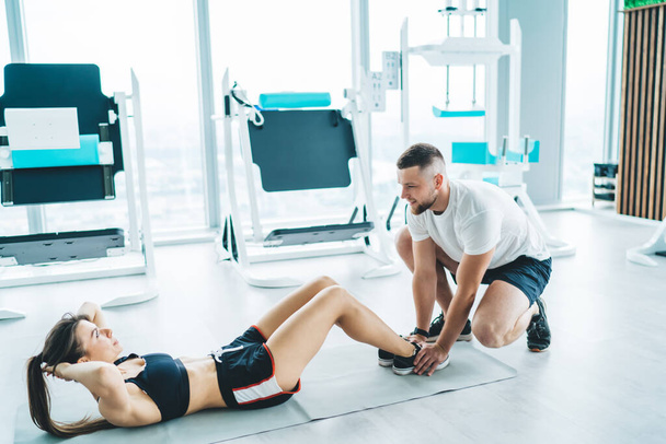 From above of full body focused woman in sportswear practicing sit ups on mat with bearded coach in modern gym - Foto, Bild