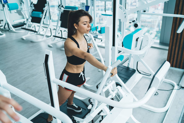 From above of full length of young woman in top and shorts training on fitness equipment in modern bright gym - Foto, Imagen