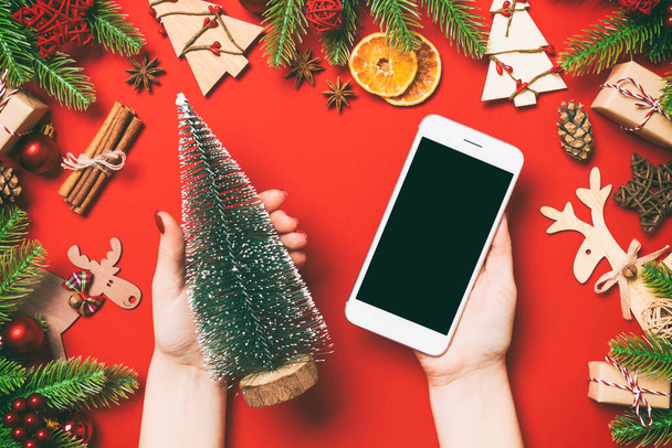 Top view of female hands holding phone in one hand and christmas tree in another hand on red background. New Year holiday concept. Mockup. - Photo, image