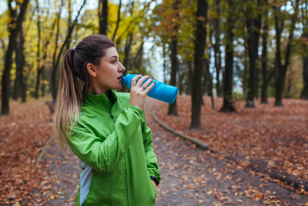 Läufer ruhen sich nach dem Training im Herbstpark aus. Frau trinkt Wasser sitzend auf dem Weg. Sportlich aktiver Lebensstil. Herbst-Workout an der frischen Luft - Foto, Bild