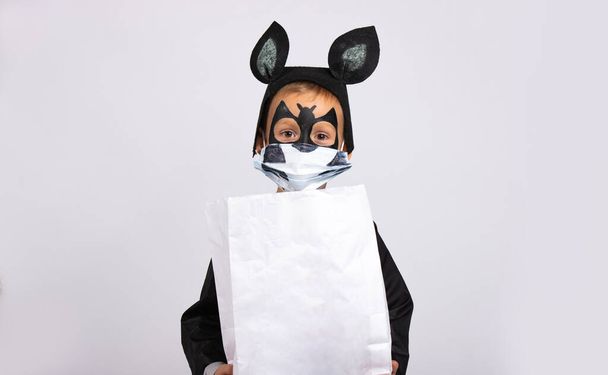  Halloween, little cute boy dressed as a bat holding a white bag of chocolates with empty space .Trick or treat. Child in a medical mask on a gray. The virus has attacked the world. High quality photo - Foto, Imagen