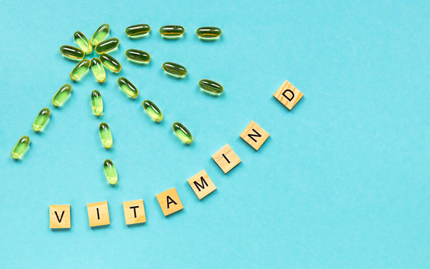 Yellow capsules in the form of the sun with rays and word vitamin D from wooden cubes with letters on blue background, healthy and medical concept.  - Foto, imagen