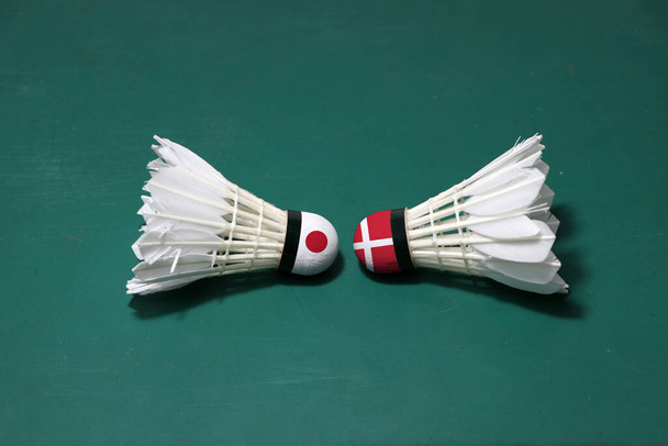 Twee gebruikte shuttlecocks op de groene vloer van Badminton Court met beide hoofden tegen elkaar. Een hoofd geschilderd met Japanse vlag en een hoofd geschilderd met de Deense vlag, concept van badminton competitie. - Foto, afbeelding