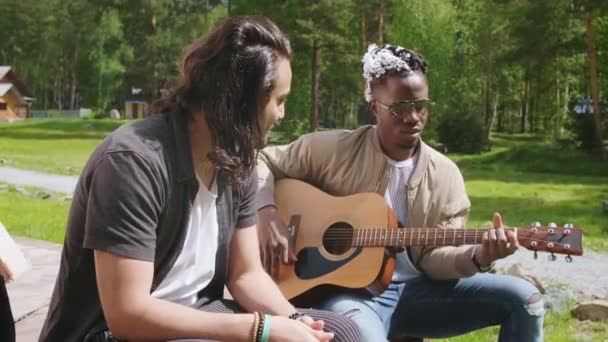 Średni materiał filmowy dwóch wieloetnicznych chłopców siedzących na zewnątrz śpiewających piosenki i grających na gitarze latem cieszących się atmosferą i relaksujących - Materiał filmowy, wideo