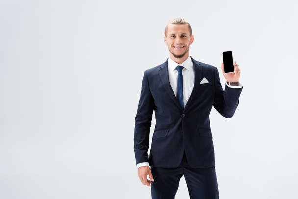 smiling young businessman in suit presenting smartphone isolated on white - Photo, Image