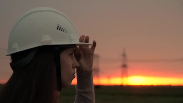 Egy gyönyörű lány mérnök fehér sisakban nézi a vezetéket naplementekor. A nő energikus az ég ellen. Villamosenergia-elosztás és -ellátás Tiszta energia. Közelkép - Felvétel, videó