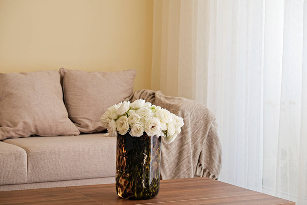 Close up shot of wooden coffee table with bouquet of beautiful white ranunculus flowers in glass vase on foreground and beige couch on the background. Copy space for text, close up. - Zdjęcie, obraz