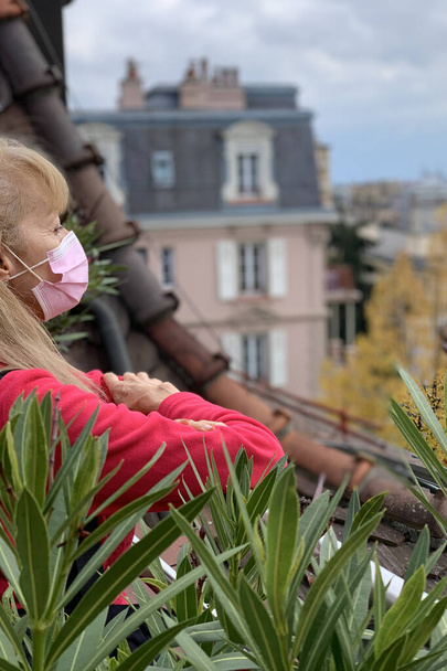 Blanke vrouw kijken vanaf het balkon in de nieuwe normaliteit, met roze chirurgische masker - Foto, afbeelding