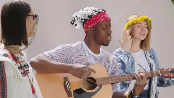 Střední detailní záběr mladých přítelkyň a afroamerického hudebníka chytání vibrace sedí mezi stany v kempu hraje na kytaru a zpěv - Záběry, video