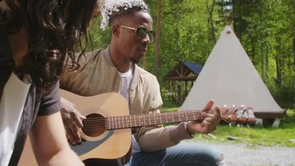 Średni materiał filmowy o mieszanej rasie długowłosego młodzieńca słuchającego swojego afroamerykańskiego muzyka z czarno-białymi dredami śpiewającego i grającego na gitarze w parku latem - Materiał filmowy, wideo