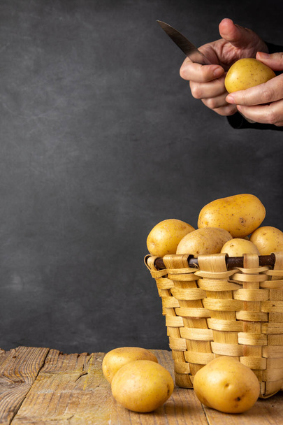 Close-up of basket with yellow potatoes and woman's hands with paring knife, on rustic table, with black background, in vertical, with copy space - Fotó, kép