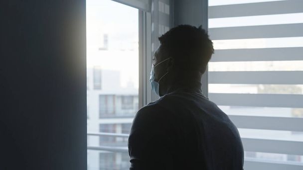 Afrikkalainen amerikkalainen musta mies lääketieteellisessä naamiossa seisoo ikkunan edessä katsomassa ulos. Karanteenin ja mielenterveyden käsite - Valokuva, kuva
