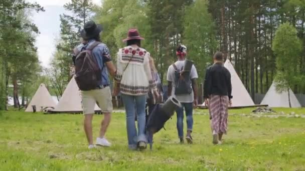 Taustakuva täynnä puukotusta trendikkäistä nuorista kantamassa reppuja, jotka saapuvat vihreälle alueelle teltat veden vieressä - Materiaali, video