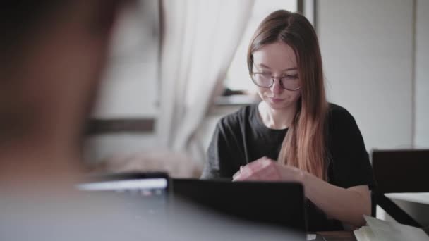 A young man and a young girl are working in front of laptops in their bright apartment. Work from home during the quarantine. Remote work of freelancers. Beautiful young pensive girl in glasses in front of a computer monitor. - Video