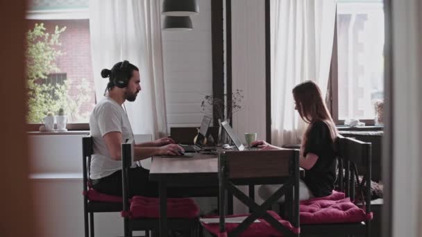 A young man and a young girl are working in front of laptops in their bright apartment. Work from home during the quarantine. Remote work of freelancers. Beautiful young pensive girl in glasses in front of a computer monitor. - Footage, Video
