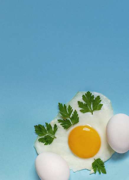 Een gebakken ei met peterselie op een blauwe achtergrond naast twee rauwe eieren. - Foto, afbeelding
