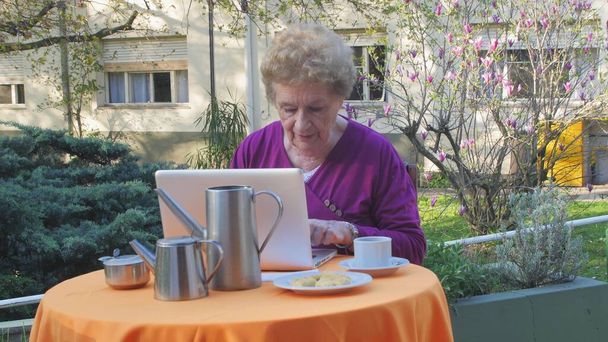 Laptop kullanan yaşlı kadın dışarıda kahvaltı hazırlıyor. Yavaş çekim. - Fotoğraf, Görsel