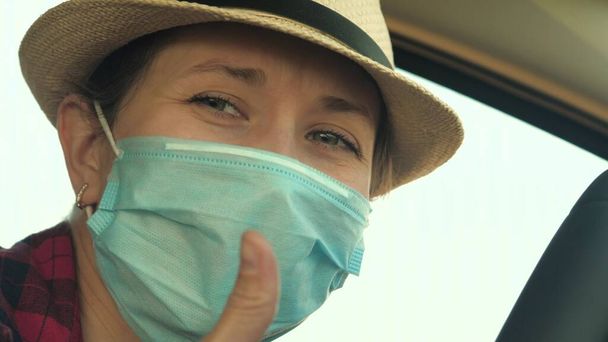 COVID-19の普及を防ぐため、医療用マスクを着用した車を運転する若い女性。車の中で顔のマスクを持つ女性の運転は、ウイルスのインフルエンザから保護する - 写真・画像