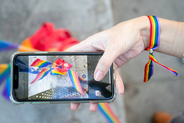 Vista dall'alto della femmina anonima delle colture nel braccialetto arcobaleno che fotografa le bandiere LGBT mentre utilizza lo smartphone sulla strada - Foto, immagini