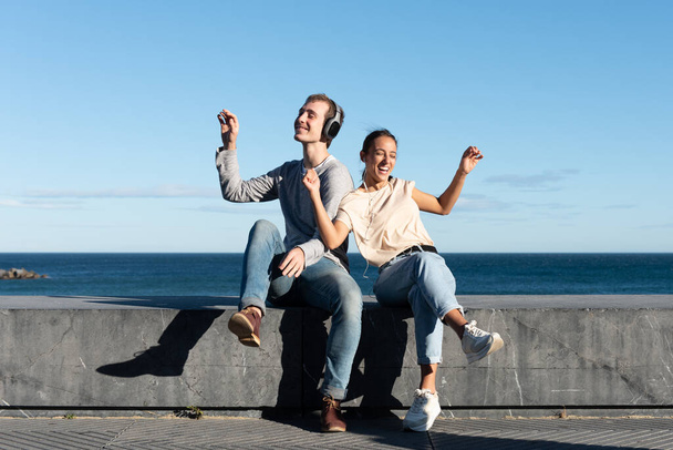 Młoda para słuchająca muzyki i tańcząca na kamiennym siedzeniu nad morzem w słoneczny dzień. - Zdjęcie, obraz