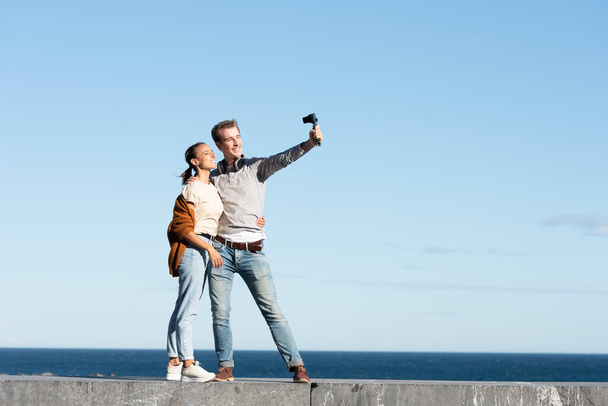 Social media content creation concept. A young couple taking themselves a selfie with a compact camera in a sunny day at the seaside. - Photo, Image