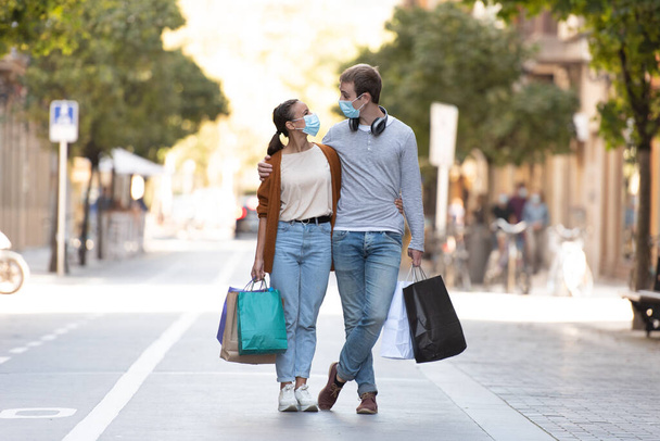 Compras durante el concepto de Coronavirus. Una joven pareja en medio de una calle peatonal con pocas bolsas de compras con máscaras médicas. - Foto, Imagen