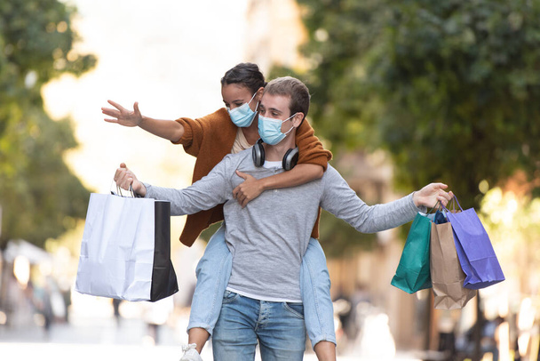 Compras durante el concepto de Coronavirus. Un hombre guapo llevando a una chica en la espalda y caminando por una calle peatonal con pocas bolsas de compras y con máscaras médicas. - Foto, imagen