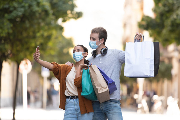 Compras durante el concepto de Coronavirus. Una joven pareja tomando una selfie con sus bolsas de compras mientras usa máscaras faciales - Foto, imagen