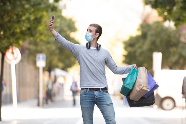 Compras durante el concepto de Coronavirus. Un joven con una máscara facial tomando una selfie mientras sostiene sus bolsas de la compra. - Foto, imagen
