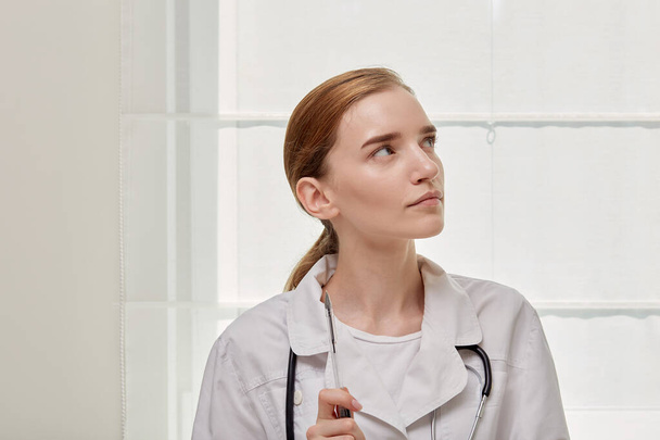 A young female doctor in a medical gown looks at the upper right corner. - Foto, Bild