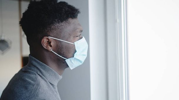 African american black man wearing medical mask standing by window looking outside. Quarantine and mental health problems - Фото, изображение