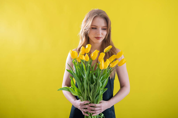 Caucasian woman with an armful of yellow tulips on a yellow background. International Womens Day. Bouquet of spring flowers - Фото, зображення