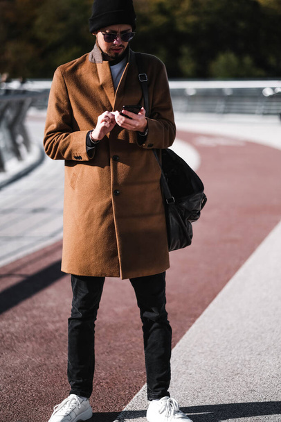Komea silmälasipäinen mies, ruskea takki ja musta hattu tekstailevat puhelimessa. muodikkaiden vaatteiden mainonta syksyllä kevät 21 / 22 - Valokuva, kuva