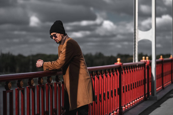 Чоловік в коричневому пальто і чорний капелюх стоїть на мосту. самотня людина, що гуляє містом
 - Фото, зображення