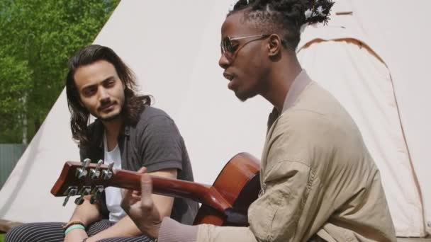 Lentitud media de jóvenes afroamericanos tocando la guitarra y cantando y la raza mixta amigo de pelo largo escuchando música acústica en verano - Imágenes, Vídeo