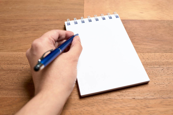 Lewa ręka młodej leworęcznej białej kobiety piszącej niebieskim długopisem na pustej kartce otwartego notesu na drewnianym biurku. Subiektywny punkt widzenia. - Zdjęcie, obraz