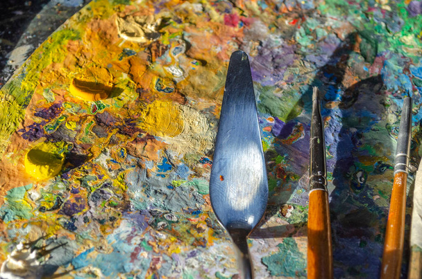 Fotografia pędzli malarskich i noża paletowego na palecie z mieszanymi i niezmieszanymi kolorami olejnymi ze światłem słonecznym - Zdjęcie, obraz