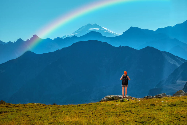 Girl traveler with backpack stands on a stone back to the viewer. She looks at the snow-capped top. Picturesque cloudless landscape with a rainbow. Wallpaper for concept of business thinking. extreme - Фото, изображение