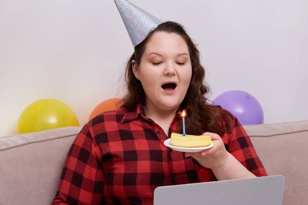 A young woman blows out the candles on a birthday cake while sitting on the sofa - 写真・画像
